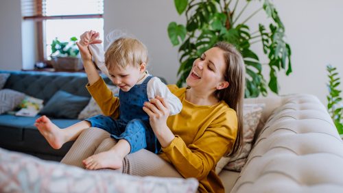 Young cheerful mother playing with her little child and having fun when lifting her up on sofa.